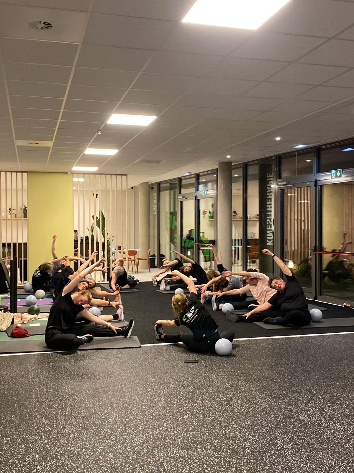 Groepsles waarbij deelnemers stretchoefeningen doen op yogamatten in fitnessruimte