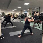 Groepsles waarbij deelnemers oefeningen doen met trainingsstokken in fitnessruimte