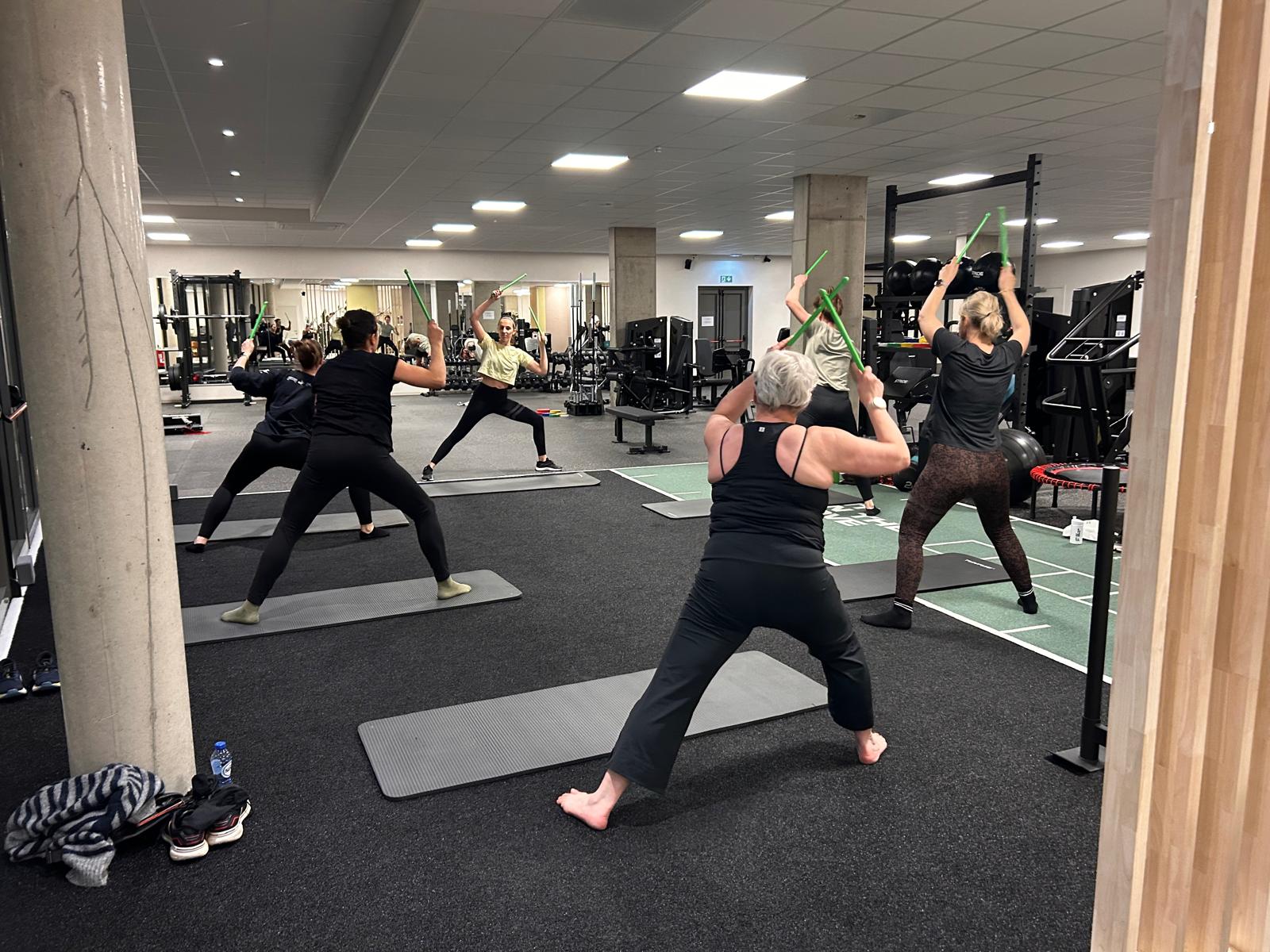 Groepsles waarbij deelnemers oefeningen doen met trainingsstokken in fitnessruimte