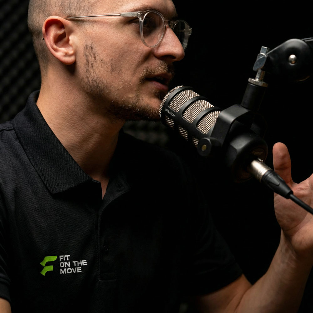 Zijaanzicht van een man met bril die in een podcaststudio in een microfoon spreekt. Hij draagt een zwarte polo met het logo “Fit On The Move” en houdt zijn hand bij de microfoon tegen een donkere achtergrond.