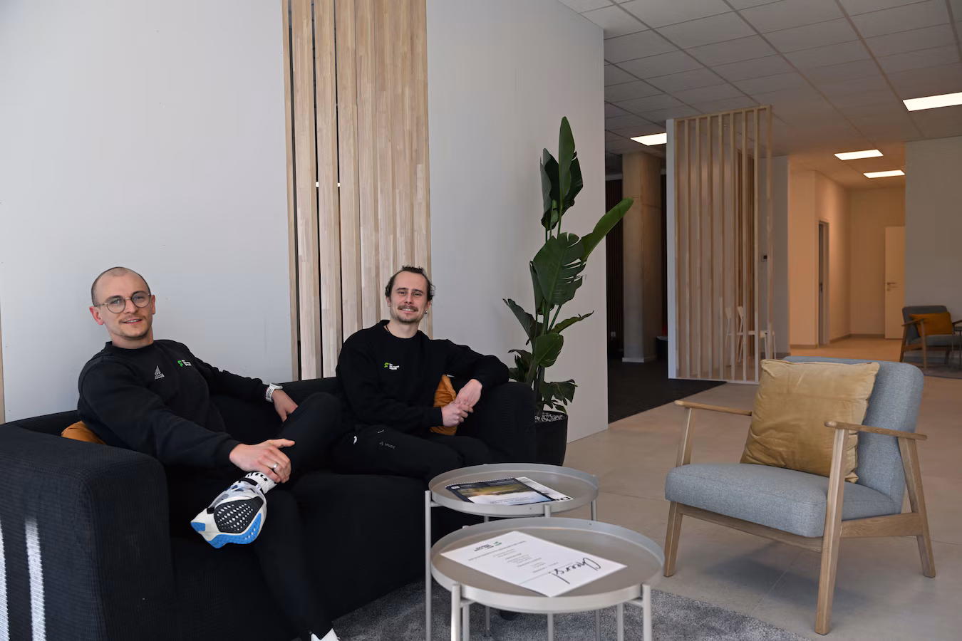 Twee mannen, Hannes Tollet en Gust Alaerts, zitten ontspannen in een moderne fitnessruimte met zitmeubilair en houten accenten tijdens de opening van hun zaak.